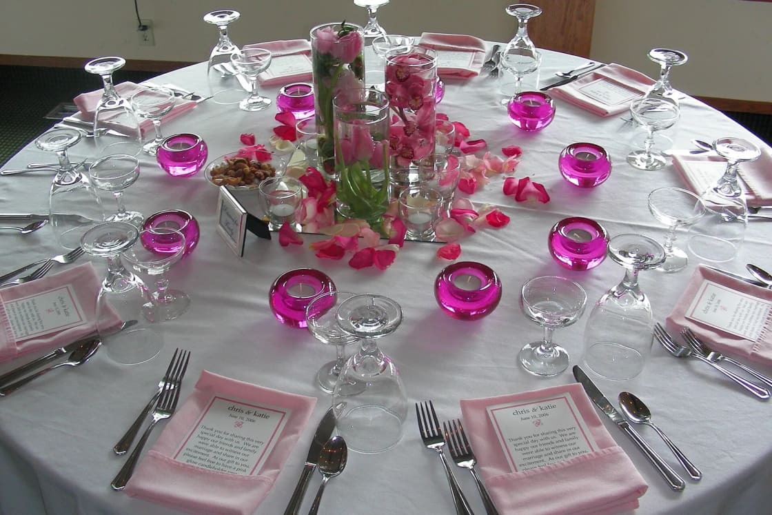 A round reception table set with pink napkins, glassware and a centerpiece of roses and candles.