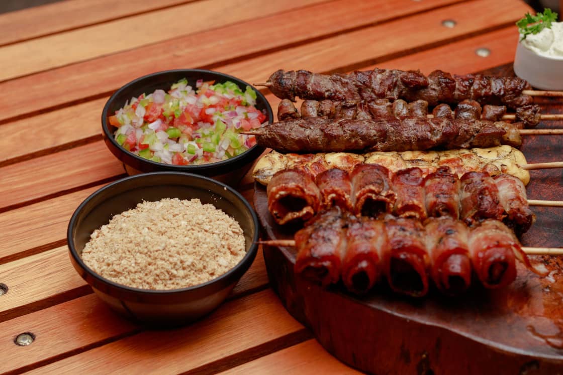 Skewers of grilled meat and bacon-wrapped bites on a wooden board beside bowls of chopped tomato-and-onion vinaigrette and toasted cassava farofa