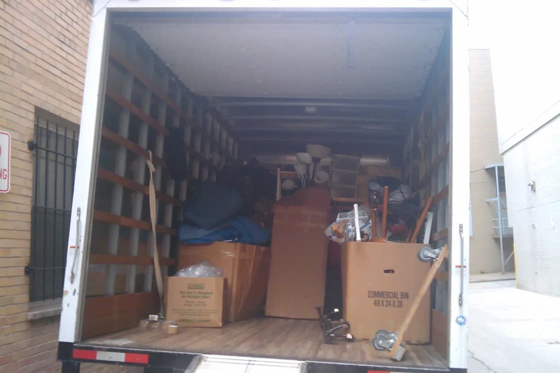 The open back of a moving truck loaded with cardboard boxes and furniture beside a brick building.