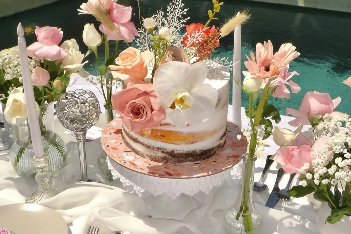 A bridal shower table set with a small layered cake, pink roses and candles, a rose-gold Bride headband resting on a white napkin.
