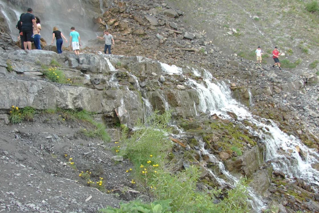 Water spilling over gray rock ledges at the base of Bridal Veil Falls, with loose rock and scree on the slope above and a few hikers in the distance