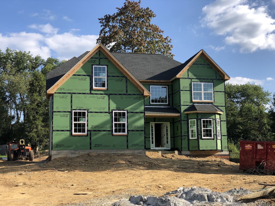 A new wood-frame house with green sheathing, roof shingles and windows installed, standing on a graded lot with a skid steer and a dumpster alongside