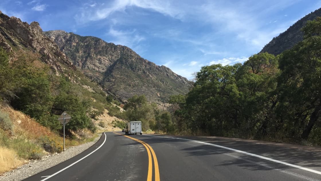 Big Cottonwood Canyon Road curving east into the canyon in Cottonwood Heights, between rocky cliffs and green scrub oak under a blue sky streaked with wispy clouds