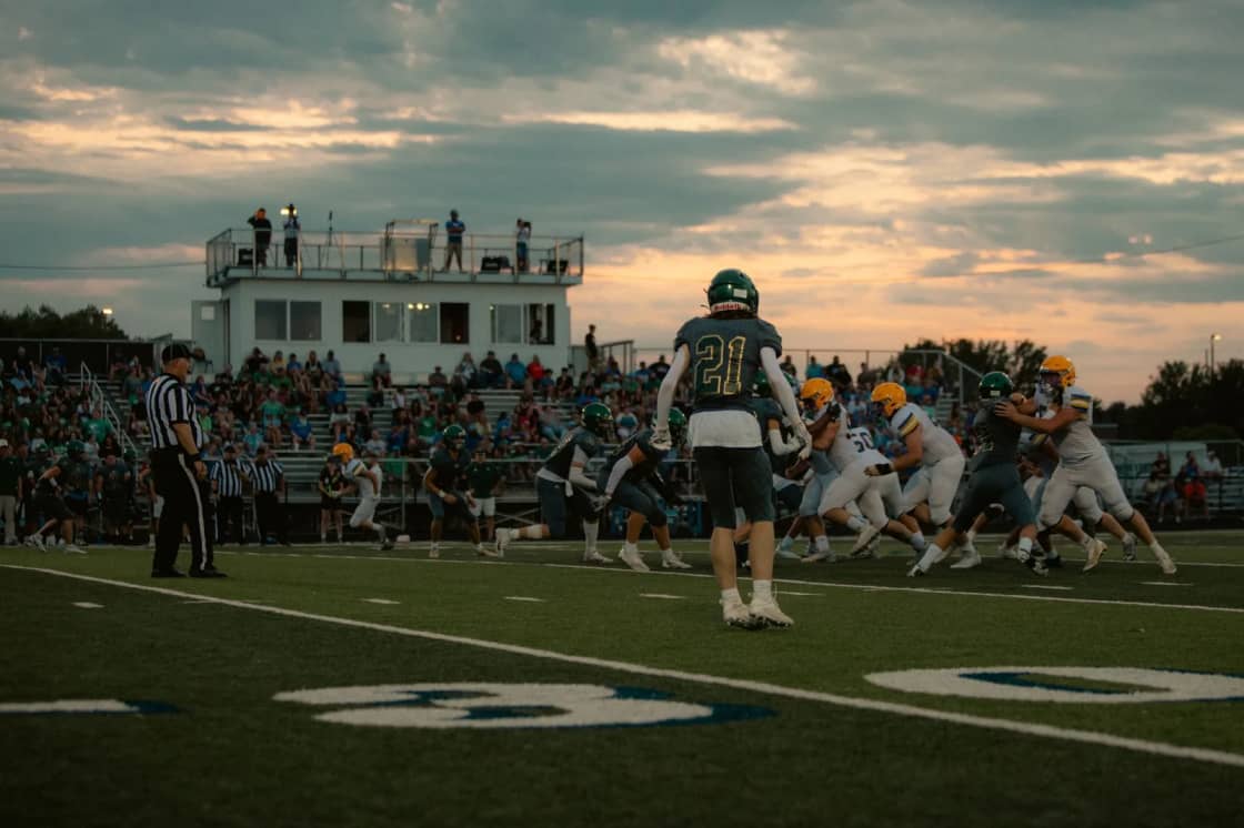 An American football game in progress under floodlights at dusk, with a referee at the left of frame and a packed grandstand behind