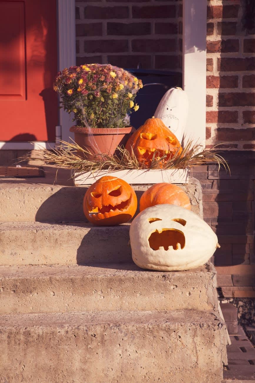 Carved jack-o'-lanterns and a white ghost-faced pumpkin arranged on concrete porch steps beside a potted chrysanthemum, a red door behind