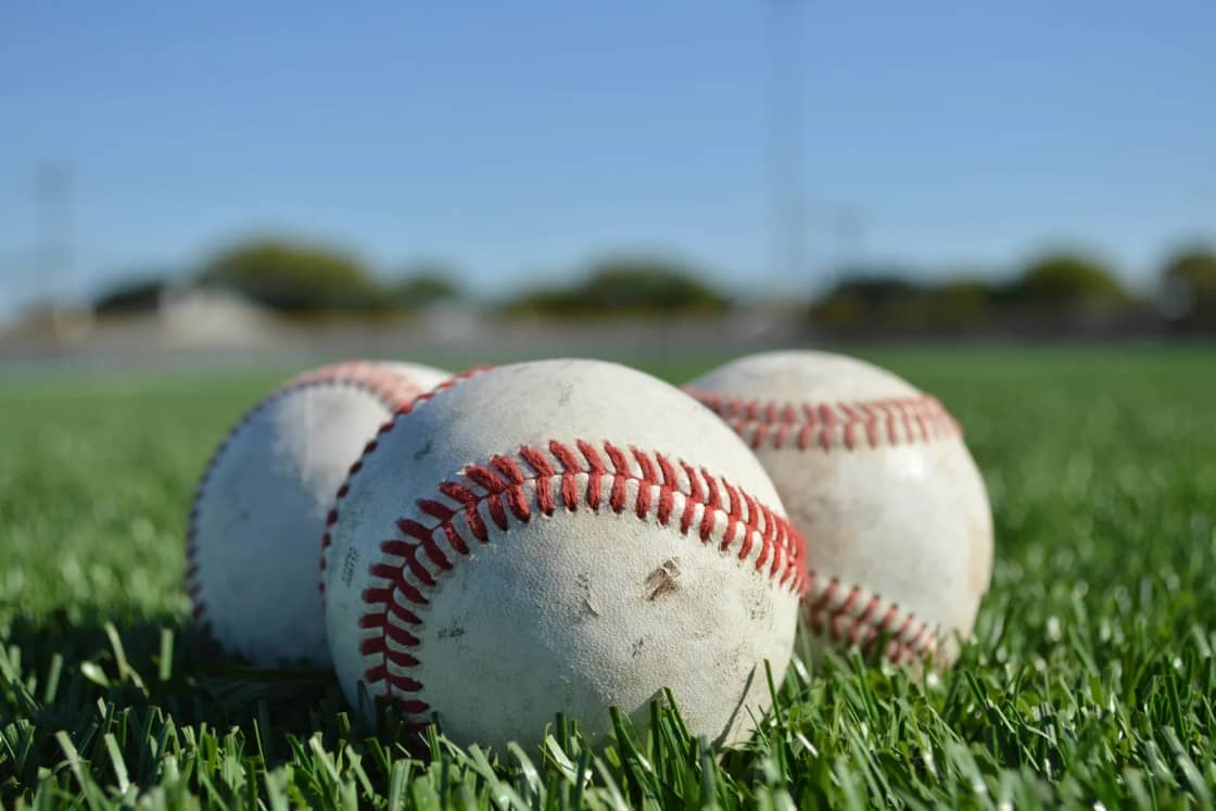 Three scuffed baseballs with red stitching resting in short green grass under a clear blue spring sky