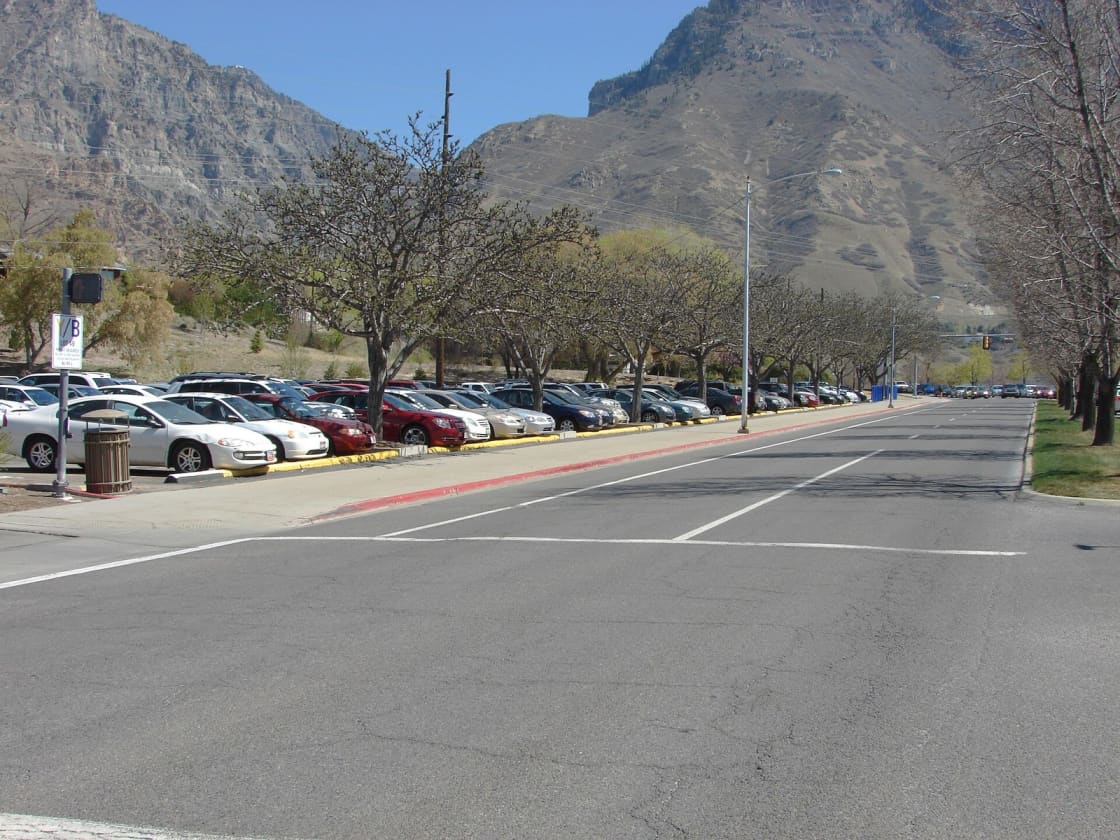 A full campus parking lot at Brigham Young University in Provo, Utah, cars nose-in along a permit-signed curb with the Wasatch Mountains rising behind