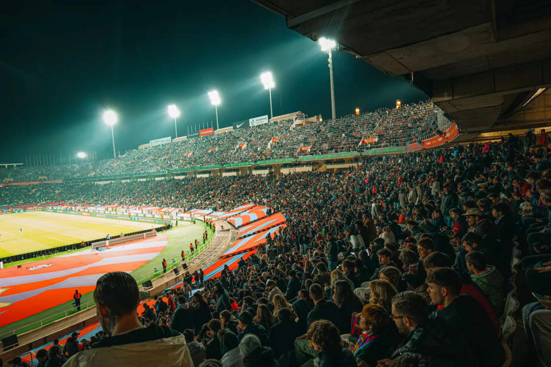A packed football stadium at night, floodlights blazing above a grandstand full of fans and a green field below — a home game during BYU Homecoming Week