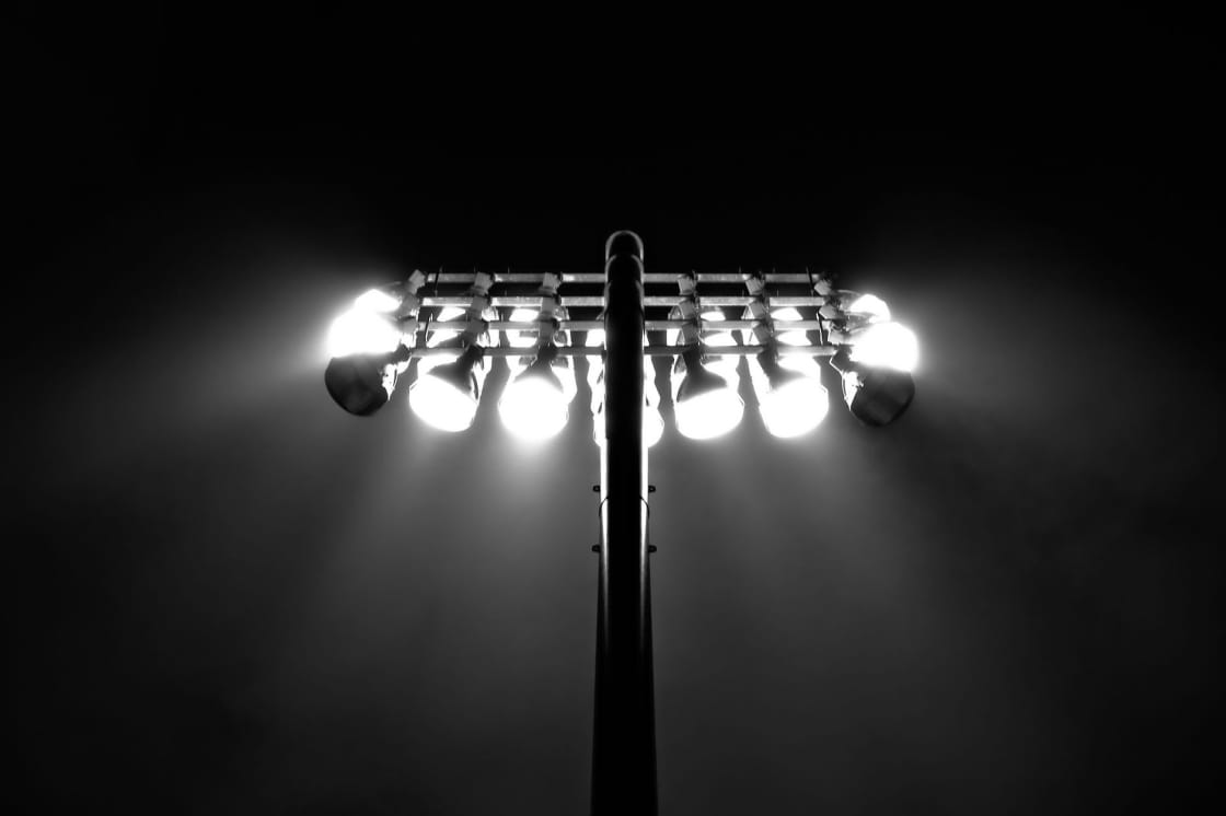 A stadium floodlight mast with two rows of bulbs burning white against a black night sky, the glare spreading in a wide halo around them