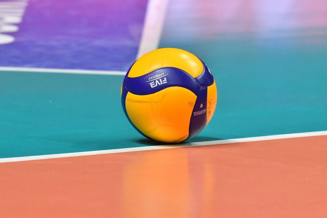 A yellow and blue match volleyball resting on the boundary line of a polished indoor court, the kind of floor the men's season is played on each winter