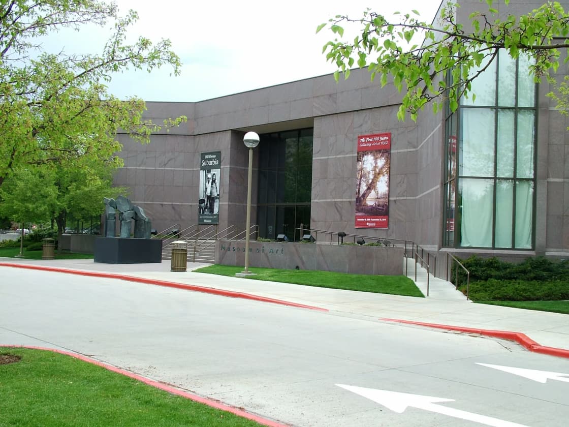 The stone exterior of the BYU Museum of Art with its name carved into the wall, two vertical exhibition banners beside the glazed entrance and a bronze sculpture group on a plinth in the plaza