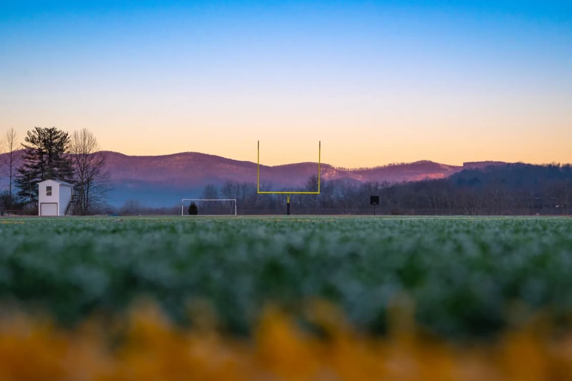 A football goalpost at the far end of a grass field at dawn, with mountains and a pink and blue sky behind