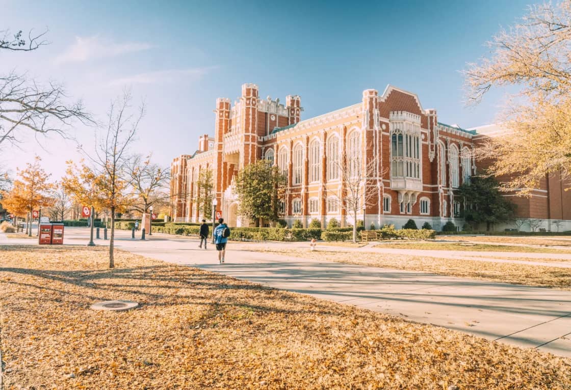 A red-brick collegiate building with tall arched windows behind bare autumn trees, with students walking on the paved plaza in front