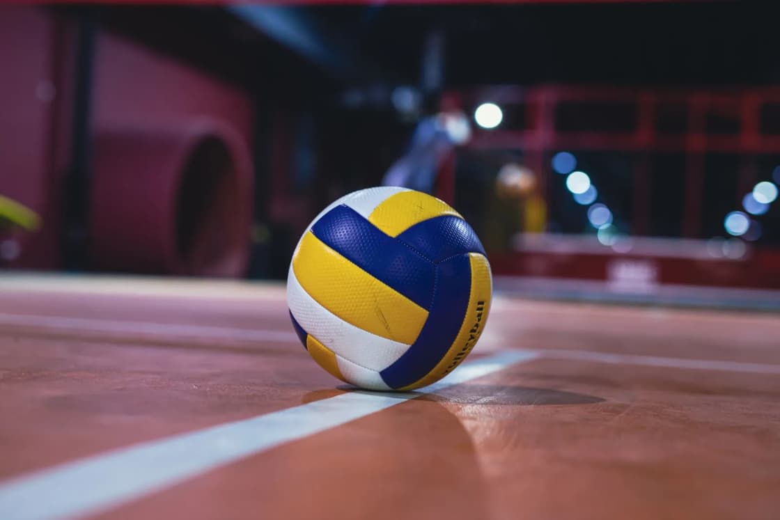 A yellow, white and blue volleyball resting on a line of an indoor court under bright arena lights