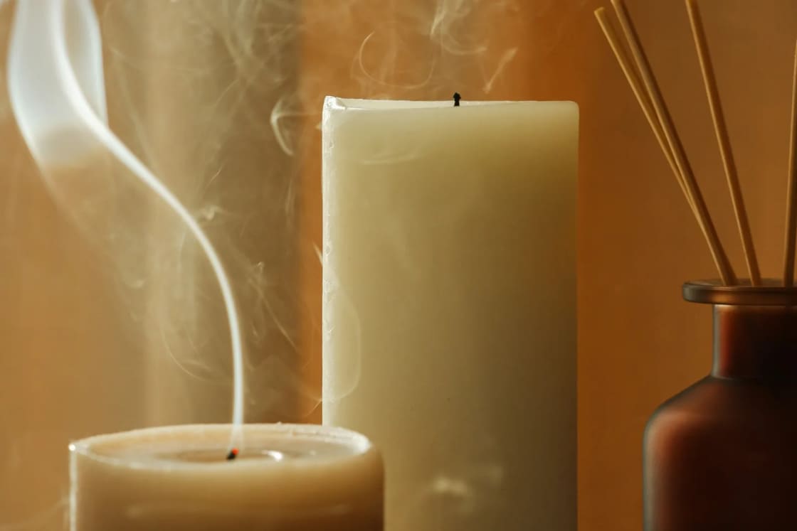 Two pillar candles, one just blown out with smoke curling up, stand beside a reed diffuser on a linen-covered table.