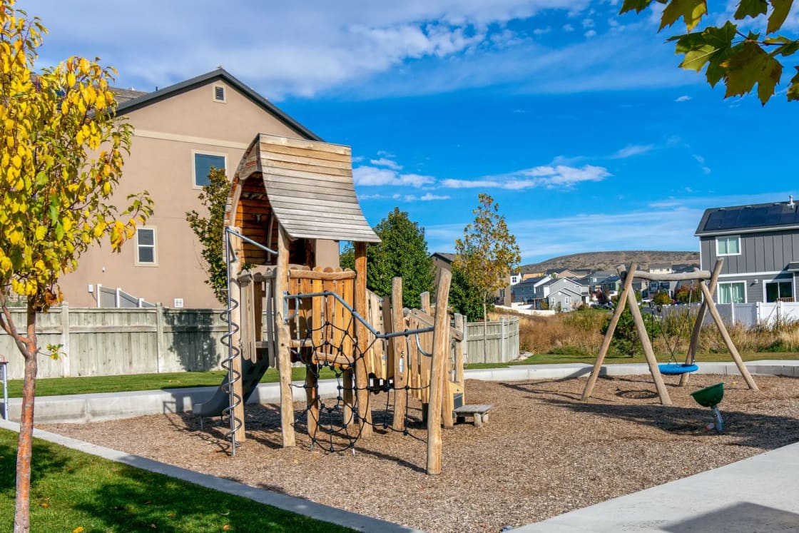 A wooden playground with a slide and climbing frame in a park beside new two-story houses under a blue sky.