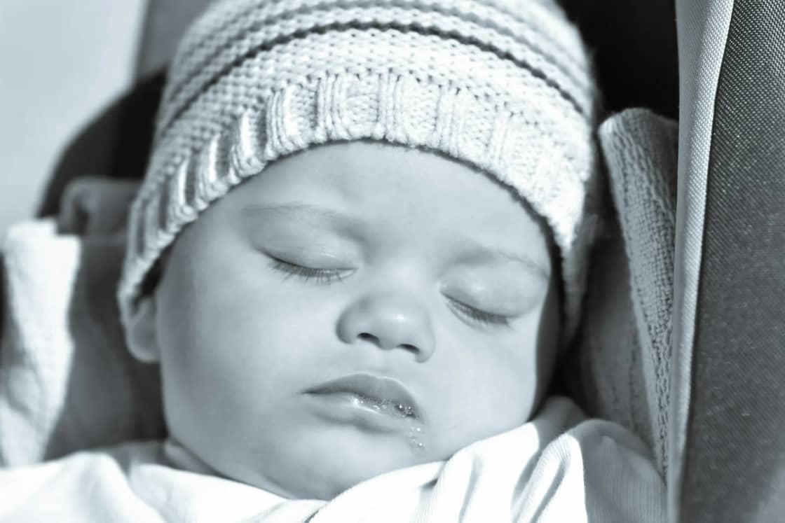 A sleeping infant in a knit cap tucked into the padded side wings of a car seat, shown in black and white.