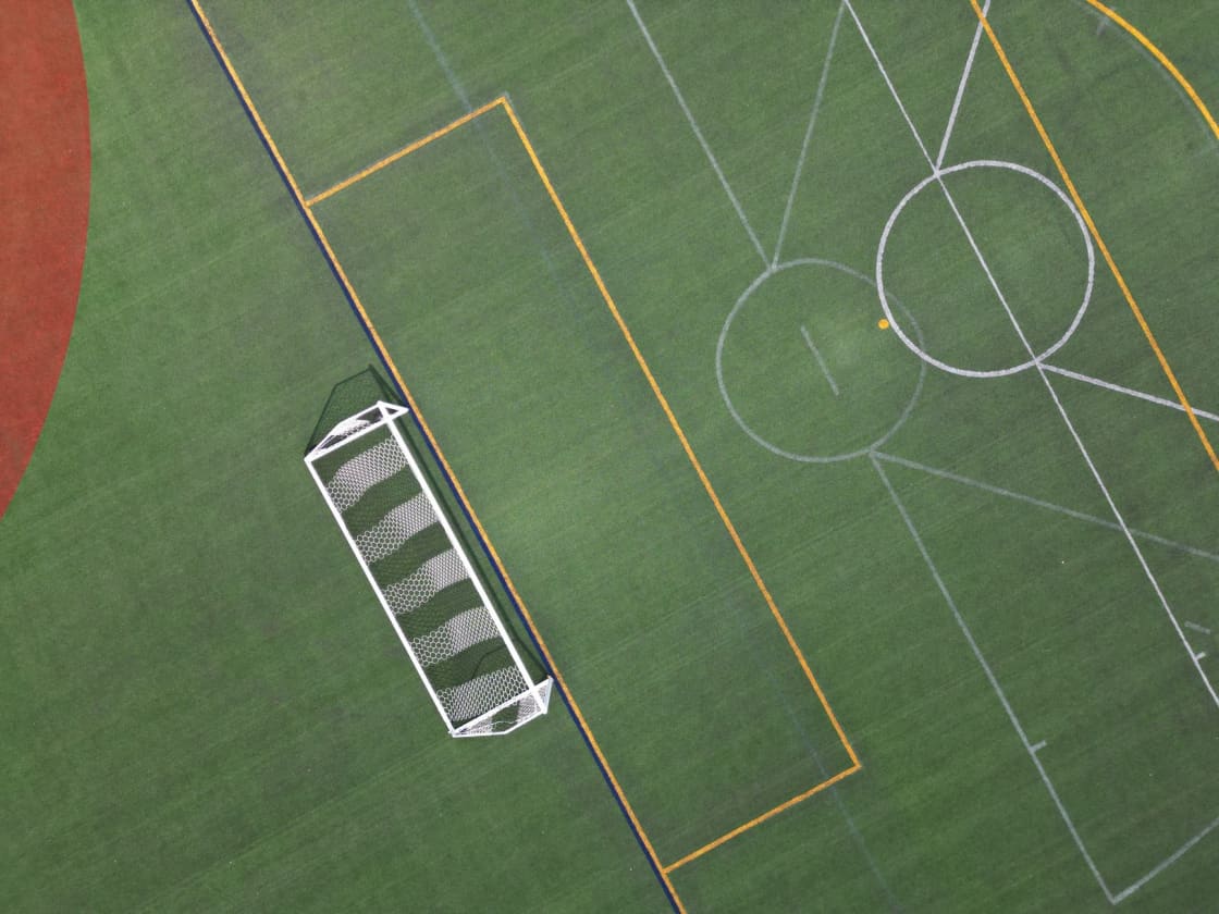 An overhead view of a green synthetic turf field with overlapping white, yellow and blue line markings, a goal frame at one edge and a red track curve in one corner