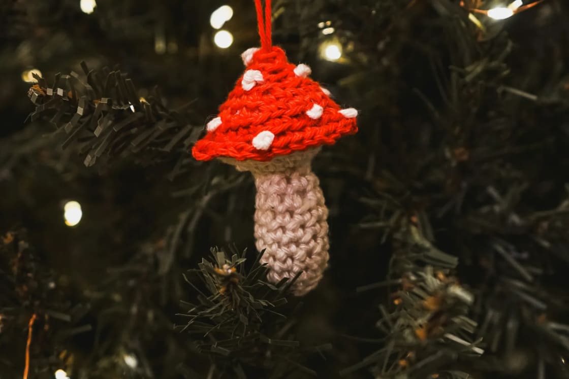 A small crocheted red-and-white mushroom ornament hangs from a red loop on a Christmas tree strung with warm lights.