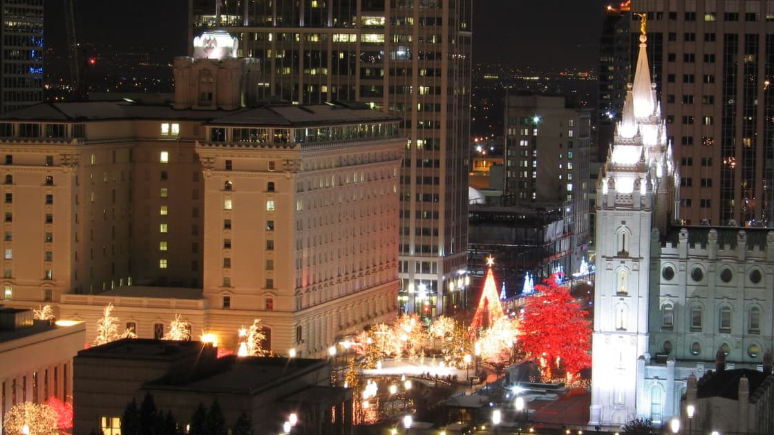 Main Street Plaza in Salt Lake City at night in December, seen from above, its trees wrapped in white and red lights between the Salt Lake Temple, the Joseph Smith Memorial Building and the downtown towers