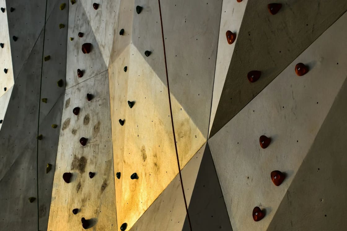 An angled indoor climbing wall studded with dark holds, a rope hanging down its face and warm light falling across the panels