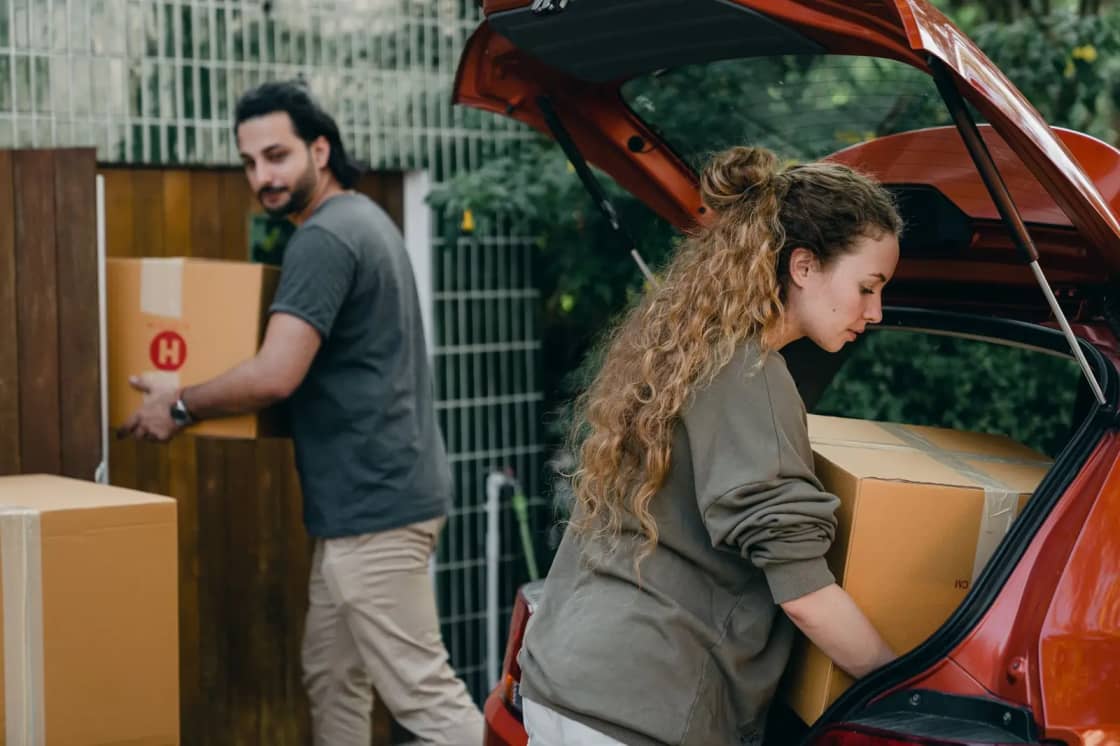 Young diverse couple in light casual clothes unloading boxes of stuff from automobile in green garden of newly purchased home
