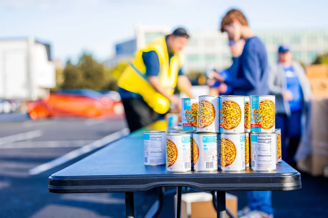 Rows of canned vegetables stacked on a folding table at an outdoor food distribution, with volunteers in safety vests working behind it