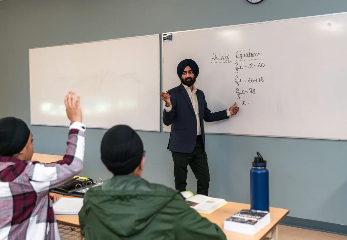A teacher in a turban writing equations on a whiteboard while adult students in a small classroom raise their hands