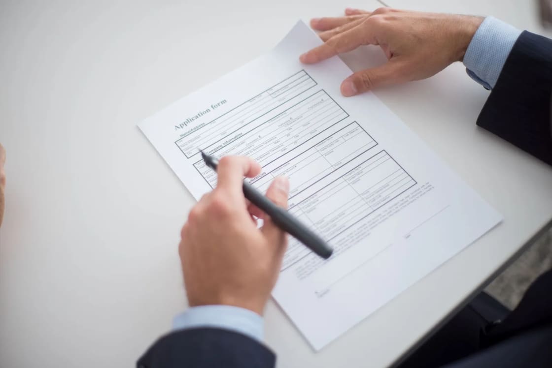 Two hands in shirt cuffs filling in a printed application form on a desk with a black pen