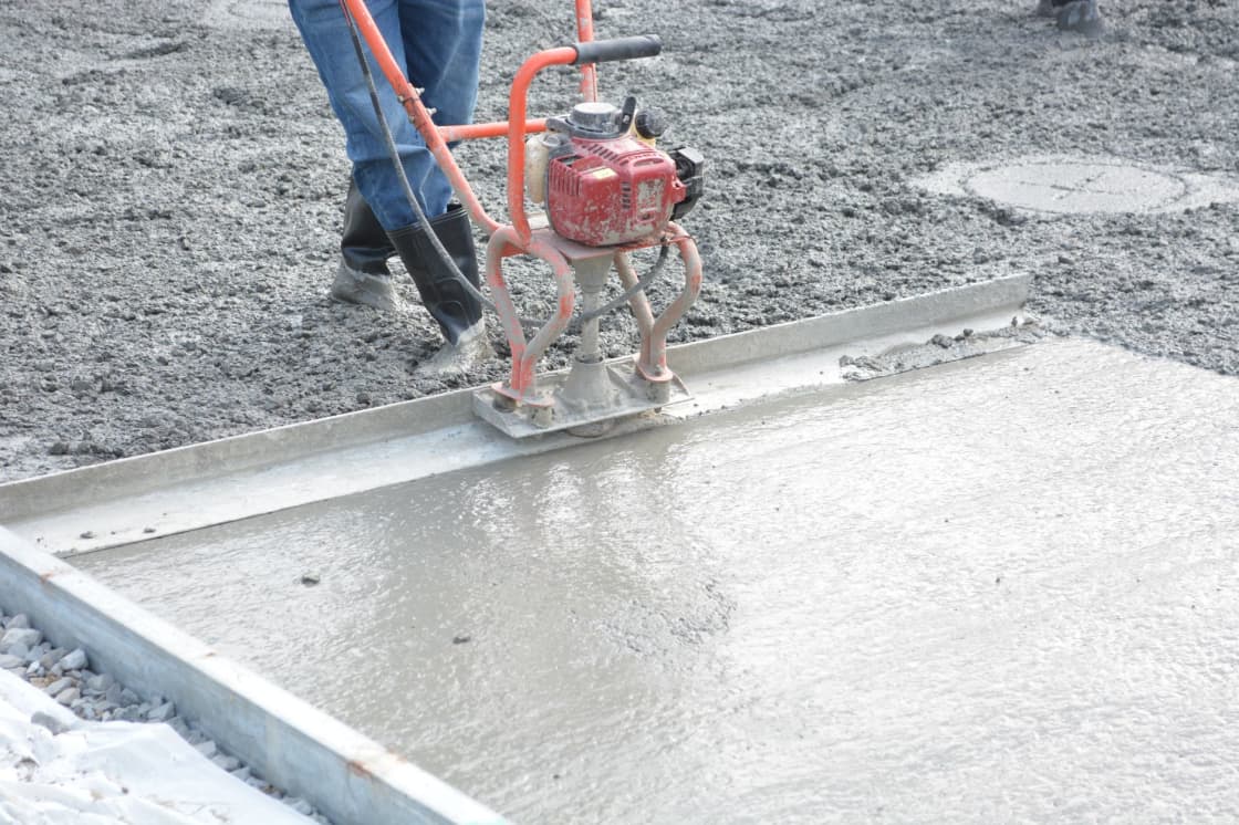 A worker running a powered screed across a freshly poured concrete slab bounded by timber forms