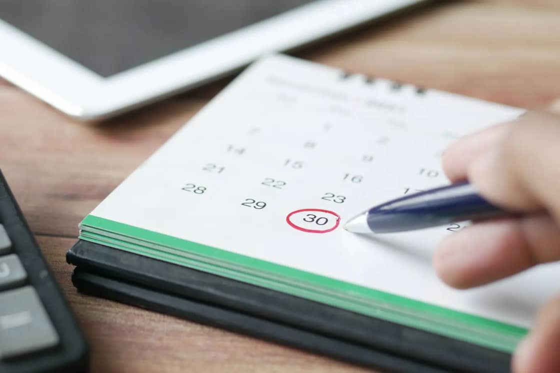 A hand circling a date in red on a desk calendar, beside a pen and a calculator