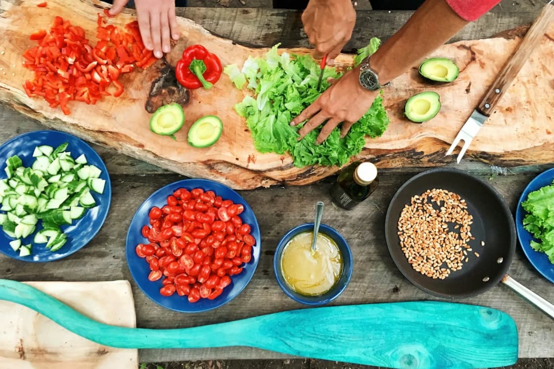Hands chopping lettuce, peppers and cucumbers on a wooden board surrounded by blue bowls of tomatoes, cucumbers, sauce and grains