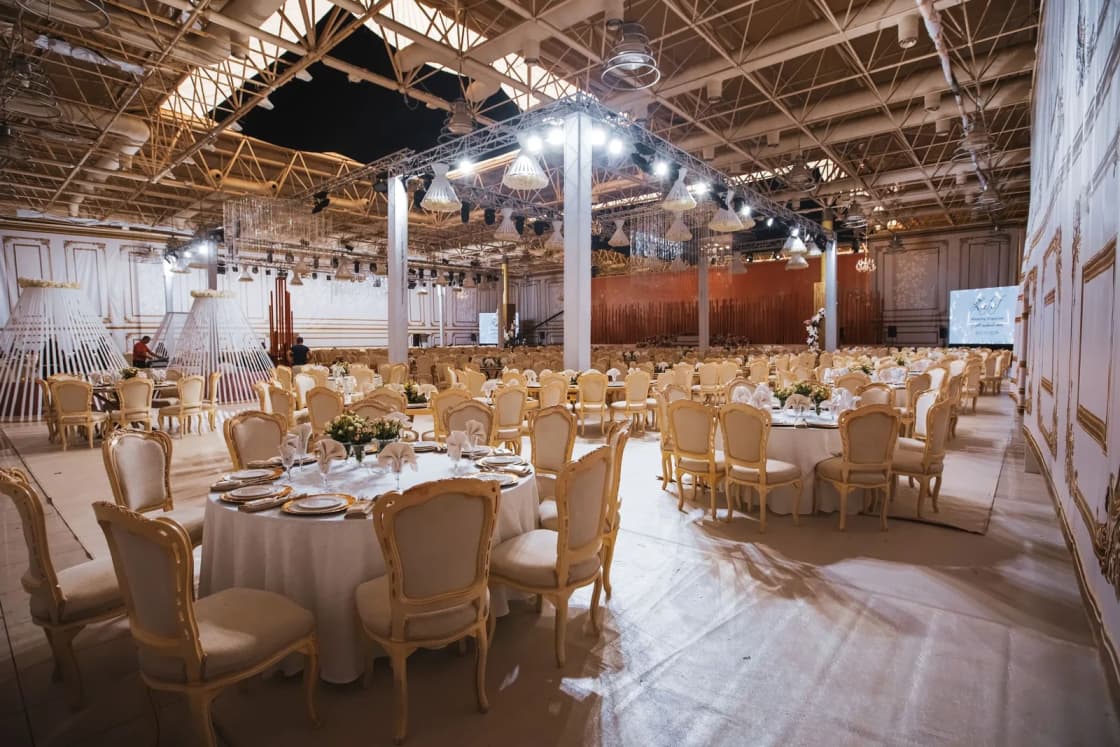 Round reception tables with white linens and gold chairs fill a large hall with exposed ceiling beams.