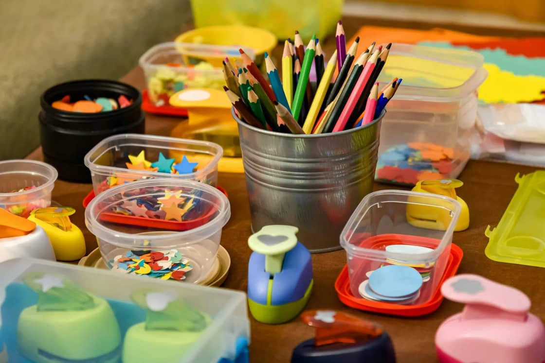 A craft table covered with colored pencils in a tin cup, paper punches and plastic tubs of cut-paper shapes ready for a group project.