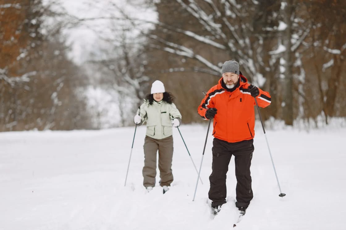 A woman in a white hat and a man in an orange jacket cross-country ski side by side along a track through a snowy forest