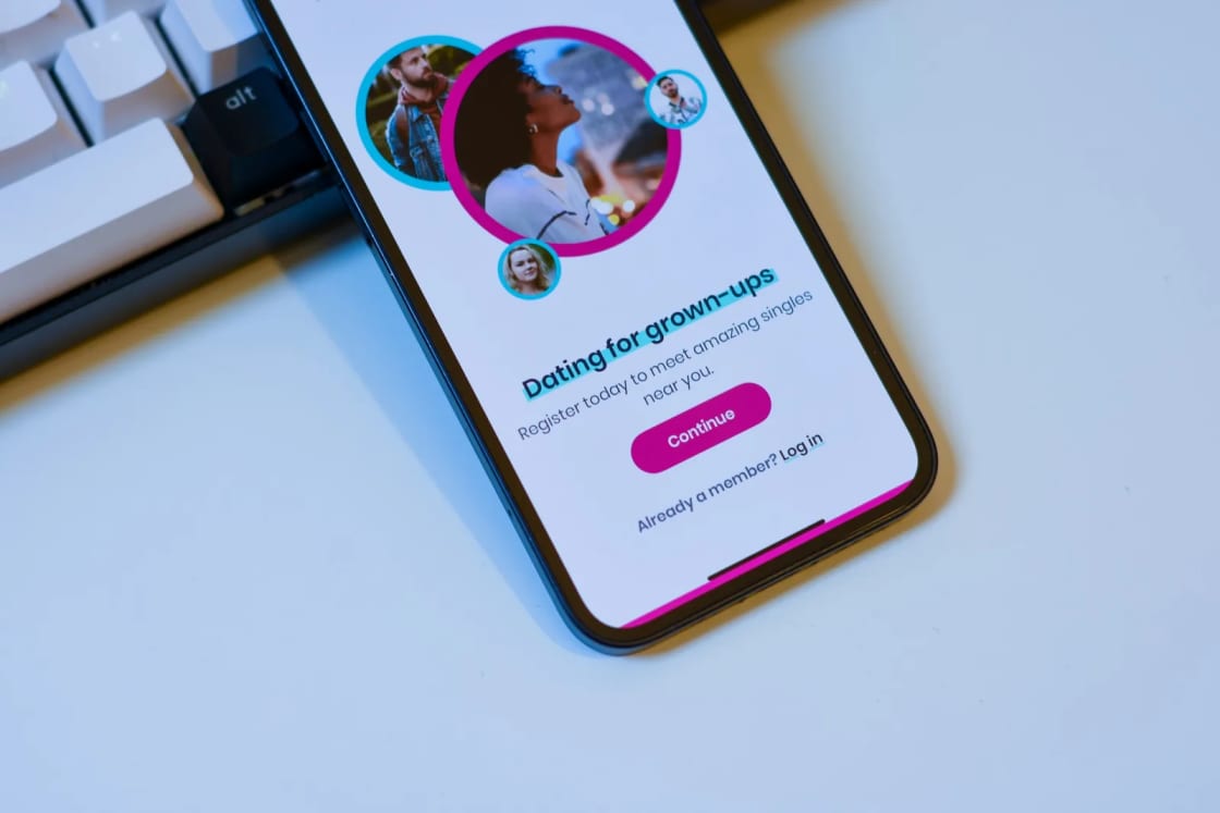 A smartphone on a white desk beside a keyboard, its screen showing the sign-up page of a dating app with profile photos