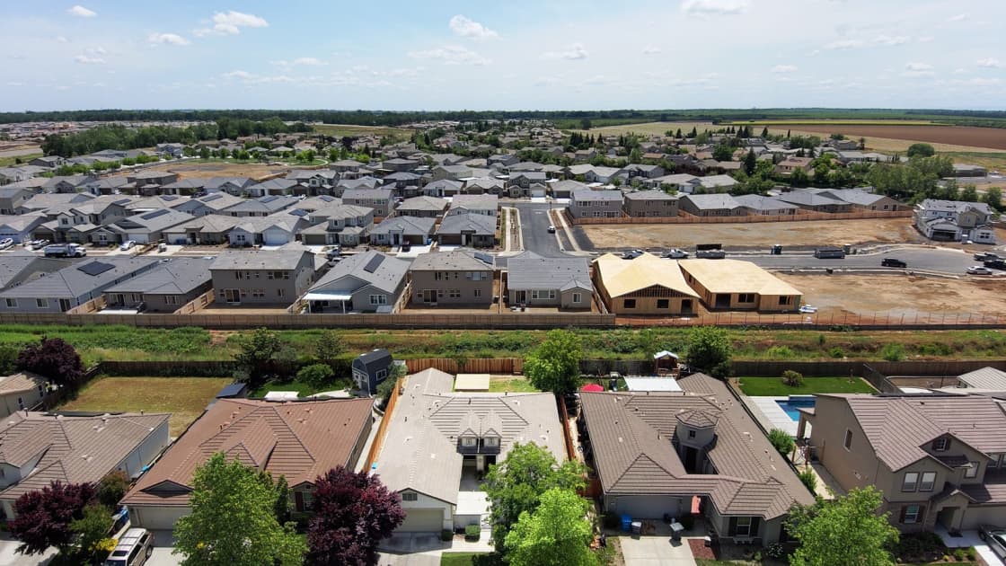 A drone view over a suburban neighborhood of new homes, with more housing under construction and open fields beyond