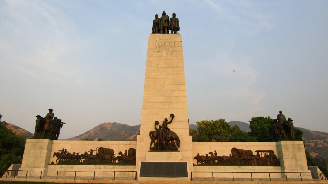 This Is the Place Monument at the mouth of Emigration Canyon in evening light, with bronze pioneer figures atop a tall stone pylon and sculpted wagon trains and horsemen along its base, the foothills behind