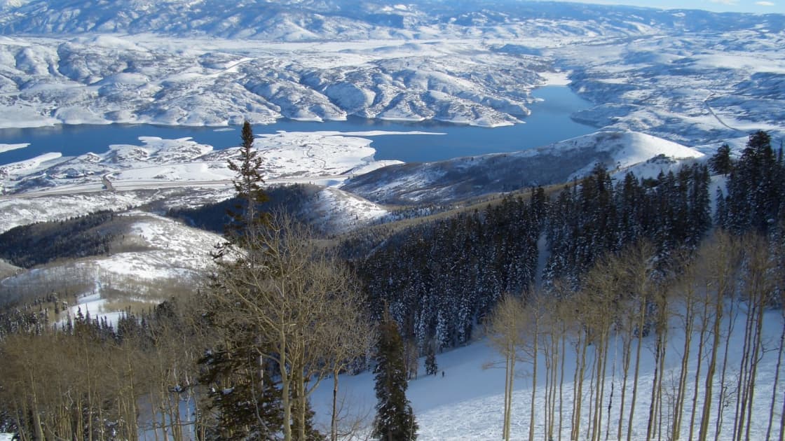 The view from a Deer Valley chairlift over snowy aspens and dark evergreens down to Jordanelle Reservoir and white rolling hills beyond, under a blue sky with thin clouds