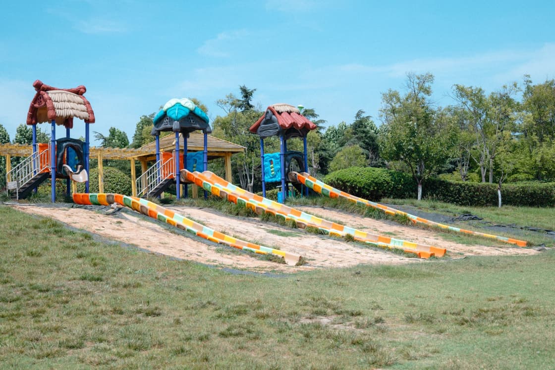 A colorful playground with long parallel slides on a grassy slope below trees