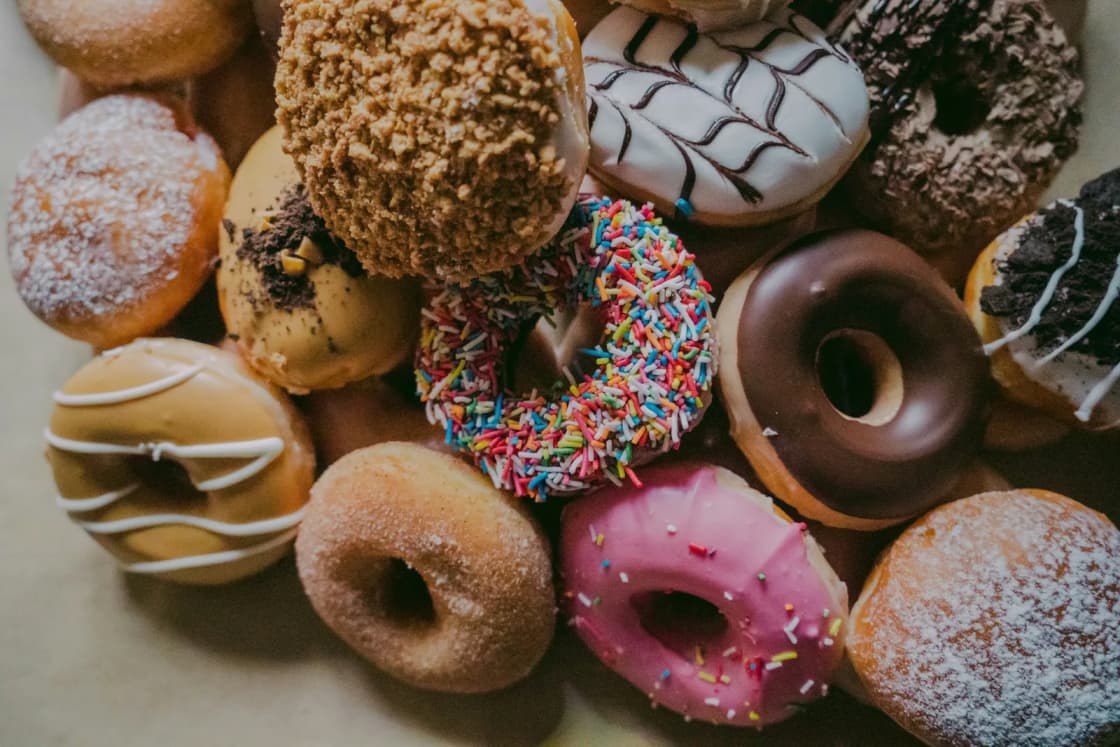 A pile of assorted donuts seen from above: chocolate glazed, pink frosted with sprinkles, powdered sugar, cookie crumble and drizzled white icing