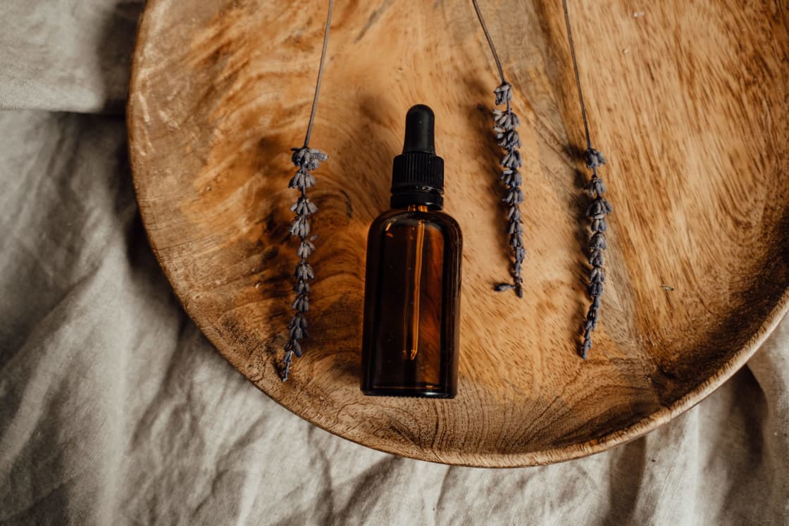 A single unlabeled amber glass dropper bottle on a round wooden platter beside three dried lavender stems