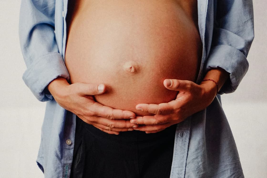 A pregnant woman in an open denim shirt cradles her bare belly with both hands, her face out of frame.