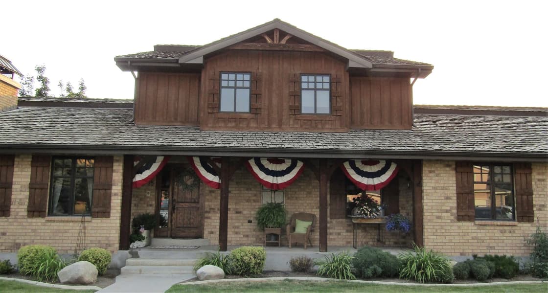 A large Utah house of brick and timber with a deep covered porch, patriotic bunting on the rails and landscaped beds along the front