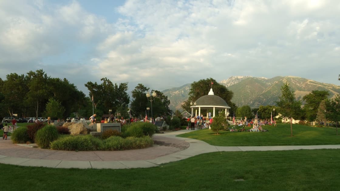 Families gather around the white domed gazebo at Draper Historic Park after the Draper Days children's parade, with decorated bikes and small flags on the lawn and the Wasatch peaks rising behind under a cloudy summer sky