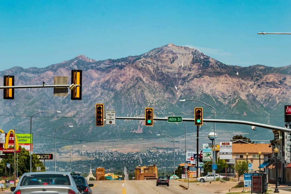 Traffic signals over a busy Wasatch Front boulevard, cars heading toward a rugged mountain under a clear blue sky
