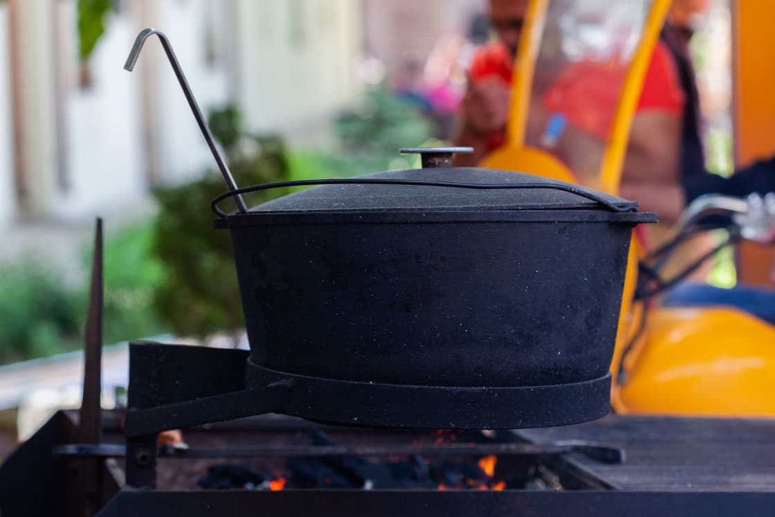 A black cast-iron camp Dutch oven with a flanged lid and wire bail handle sitting over glowing coals, a lid-lifter hook resting against the rim