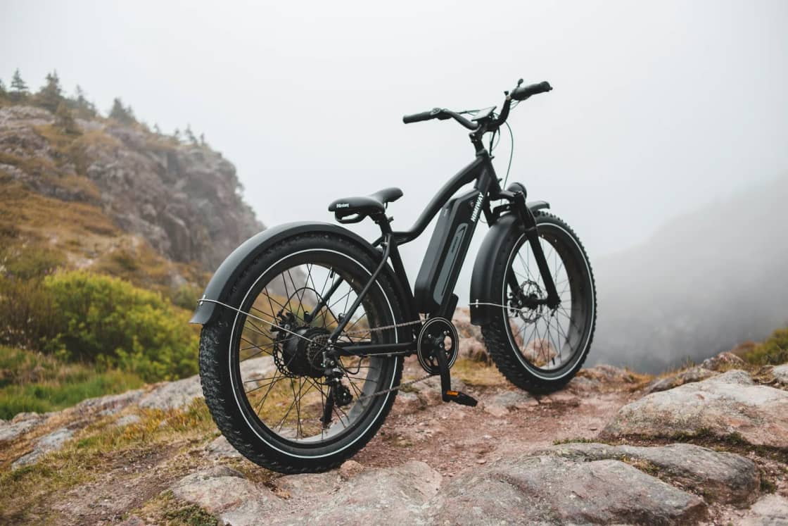 A black fat-tire electric bike with a frame battery parked on a rocky outcrop, with a misty hillside and scrub behind it