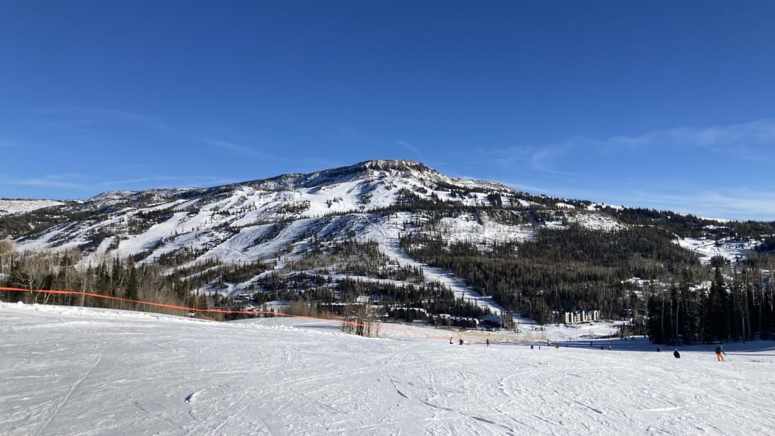Brian Head's snowy peak and ski runs under a deep blue sky, seen from a wide groomed slope with a few distant skiers