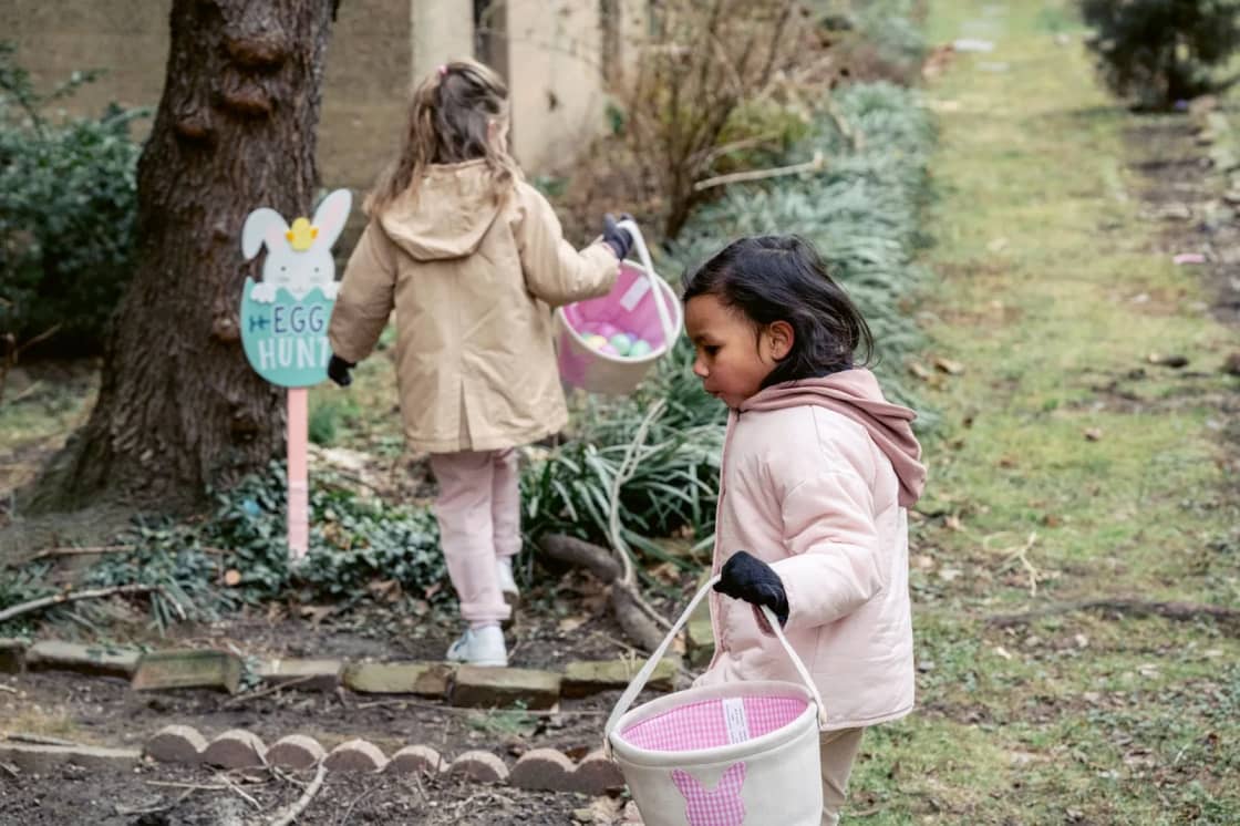 Two young girls in winter coats and gloves carry pink-lined baskets down a garden path past a wooden Egg Hunt sign, one looking down for eggs